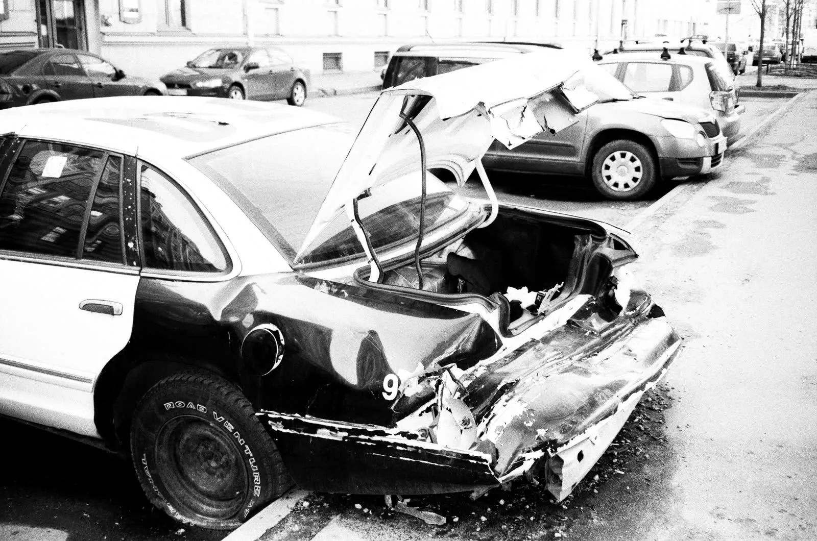 A black and white photograph of a damaged car parked on a street.